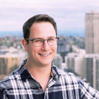 Man smiling, wearing glasses and a plaid shirt, with a blurred city skyline in the background.