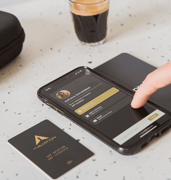 Hand interacting with a smartphone showing a profile screen, next to a black Shy Aviation card and a glass of coffee.