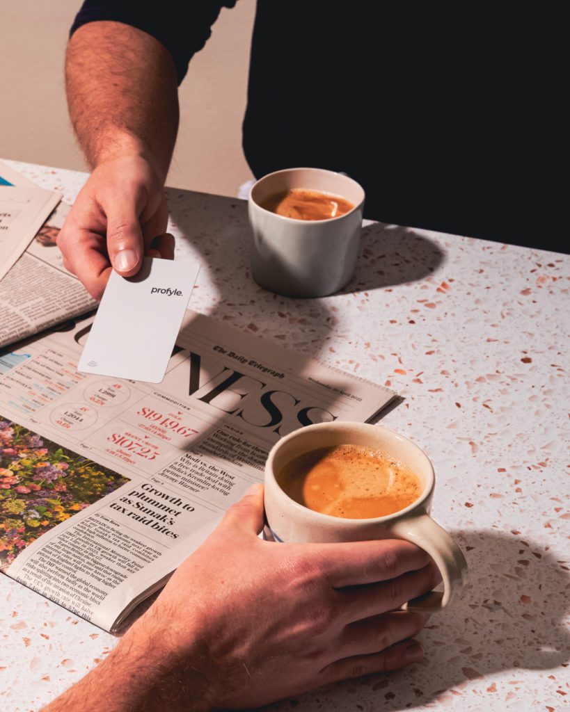 A person's hands, one holding a white Profyle card, the other a coffee mug, on a newspaper.