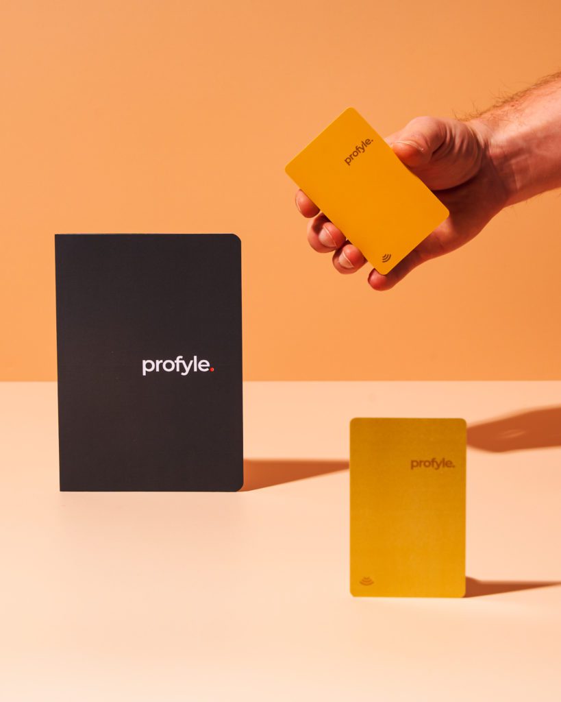 Hand holding a yellow Profyle card, with a black Profyle notebook and another yellow Profyle card on a surface.