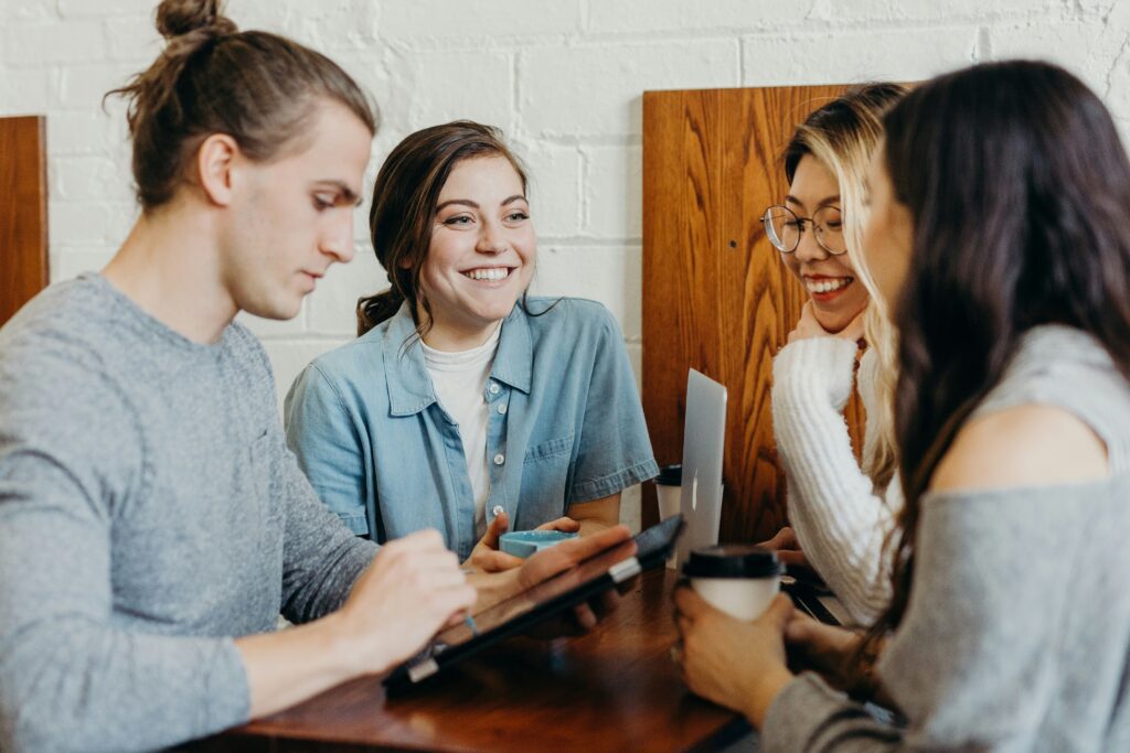 Four young adults at a table, two smiling, one using a tablet, another holding a coffee cup. A laptop is on the table.