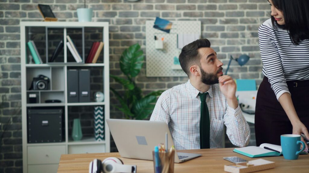 Man in plaid shirt and green tie looks up while woman in striped shirt stands beside him at office desk.