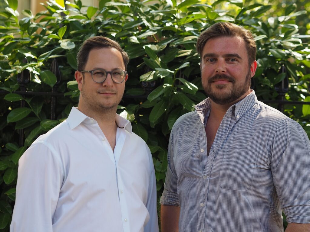 Two men stand side-by-side outdoors. One wears glasses and a white shirt, the other a beard and checkered shirt.