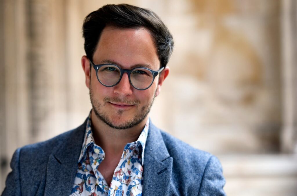 Man with dark hair, beard, and glasses smiling, wearing a blue patterned shirt and tweed jacket