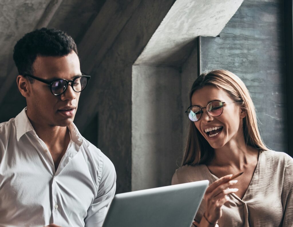 Man and woman in glasses smiling and looking at a laptop, woman holding a pen.