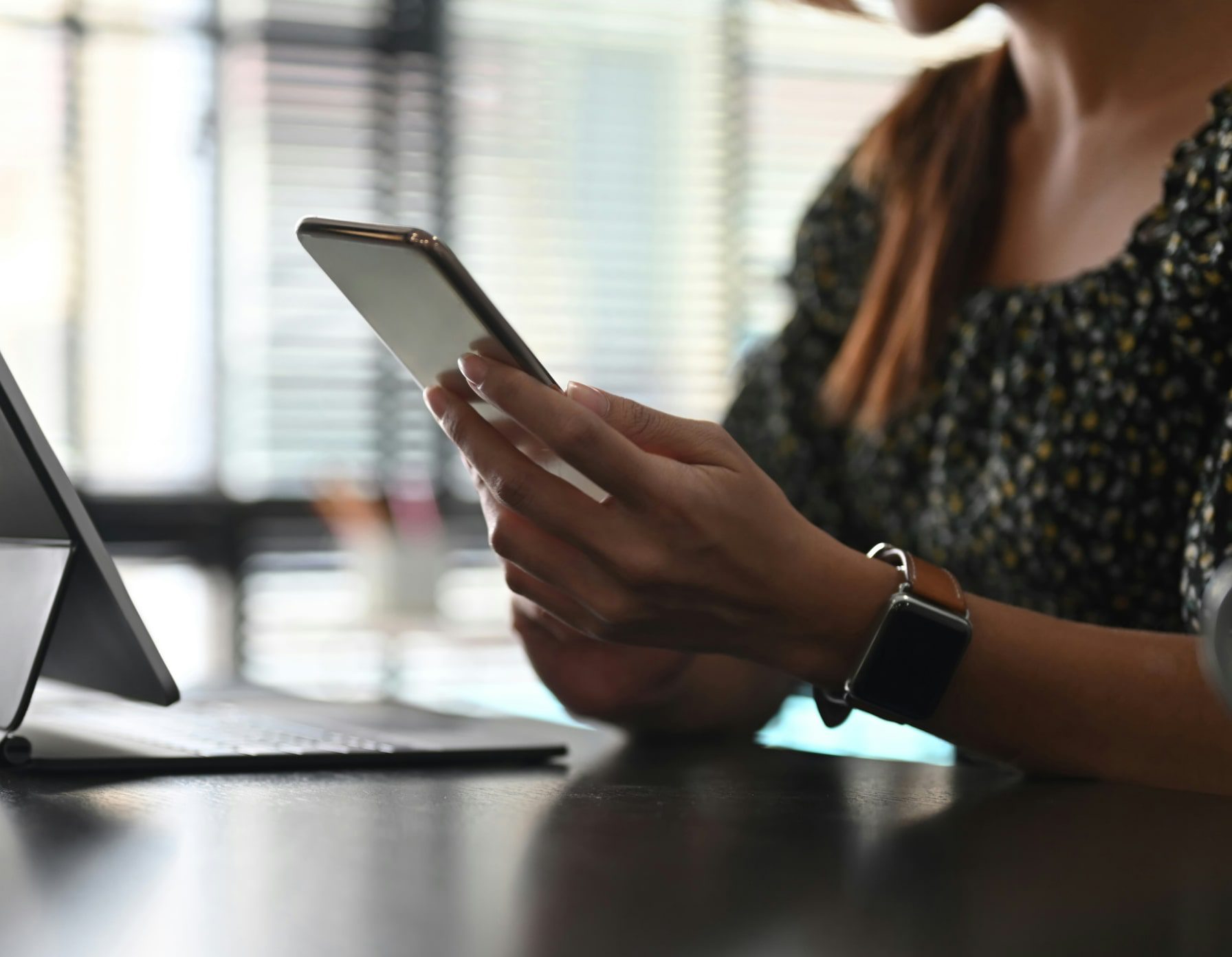 Person holding a smartphone, wearing a smartwatch, with a laptop on a desk.