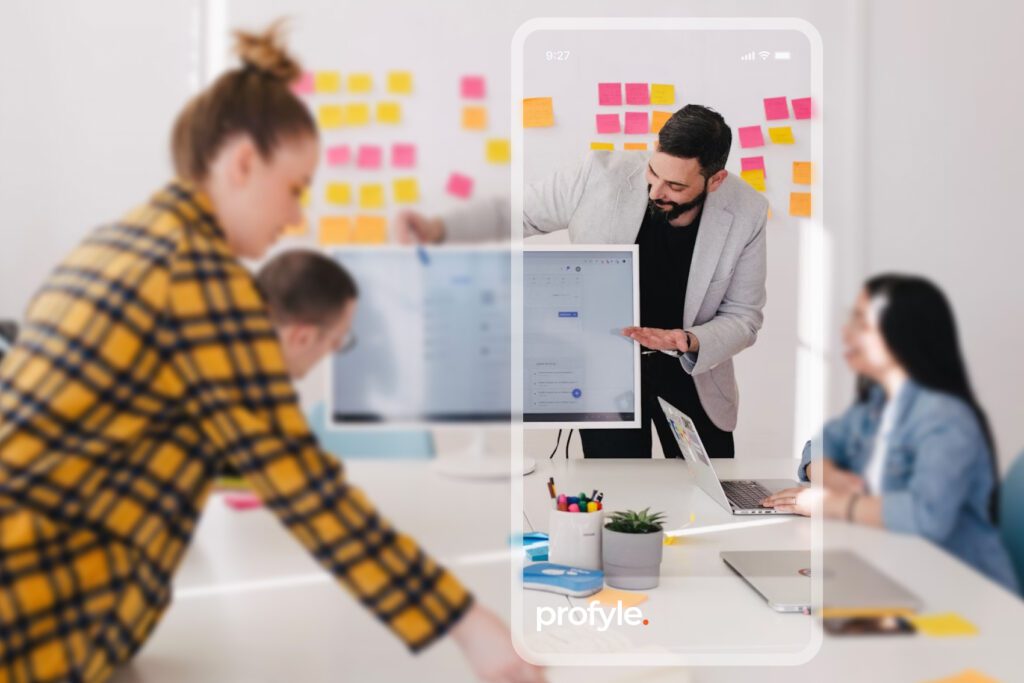 A man points at a screen displaying a user interface, with "profyle." visible, while others work in a blurred office setting.