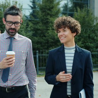 Man in glasses and woman in blazer walking outdoors, man holding coffee, woman smiling.