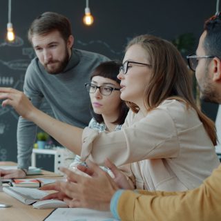 Four professionals collaborating in a meeting. A woman points and speaks while others listen.