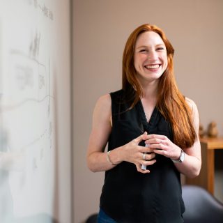 Smiling woman with red hair standing next to a whiteboard, holding a marker