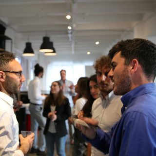 Two men talking at an indoor event, with other people in the blurred background.