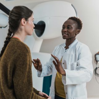 A healthcare professional in a white coat talks to a patient in a medical room with a large machine.