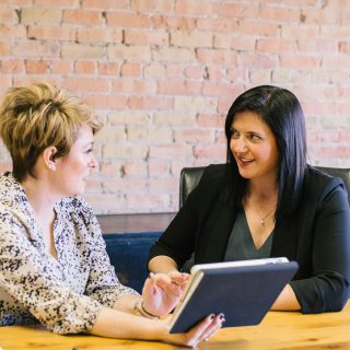 Two women discuss content on a tablet at a wooden table with a brick wall background.