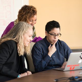 Three people looking at a laptop screen, two smiling, one with a hand on their chin.
