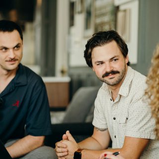 Two men with mustaches and beards, one in a patterned shirt, the other in a dark polo, engaged in conversation.