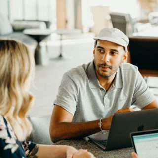 Man in a cap looking at a woman during a meeting, with a laptop open